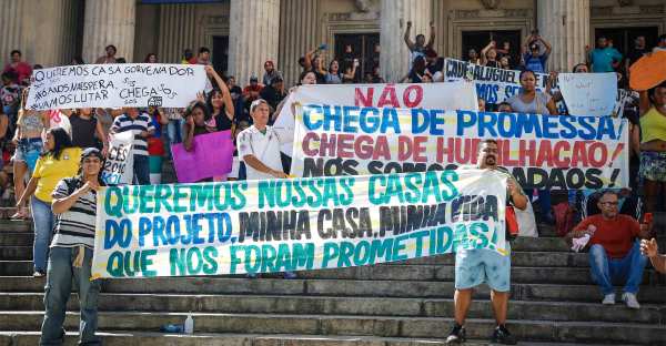 Brazilian protesters demonstrate on the steps of Rio's Legislative Assembly holding handmade signs. The sign in front reads "Queremos nossas casas do projeto, minha casa, minha vida, que nos foram prometidas!"