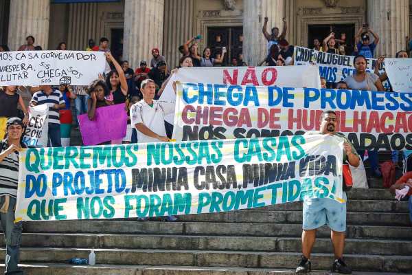 Brazilian protesters demonstrate on the steps of Rio's Legislative Assembly holding handmade signs. The sign in front reads "Queremos nossas casas do projeto, minha casa, minha vida, que nos foram prometidas!"