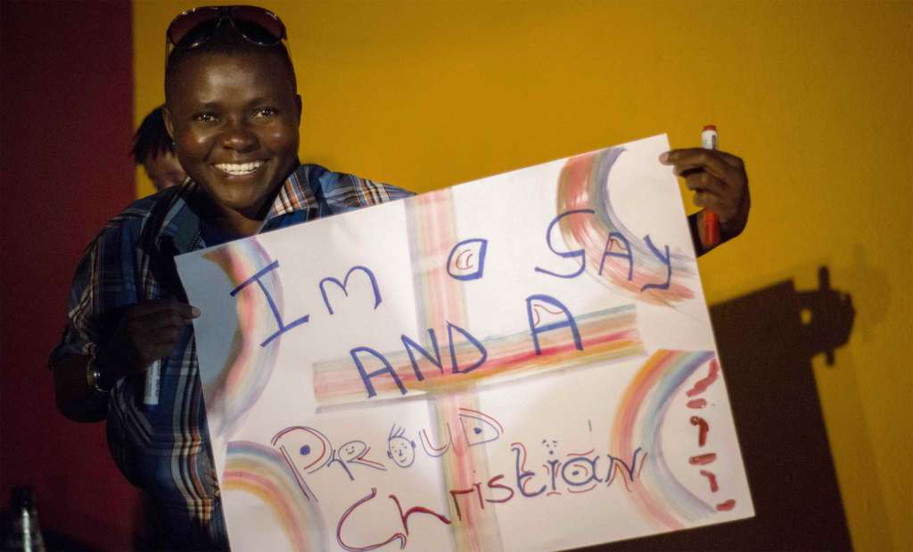 A smiling young Black man wearing a blue plaid shirt holds up a homemade protest sign that reads "I'm a Gay and a Proud Christian."