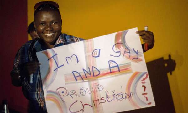 A smiling young Black man wearing a blue plaid shirt holds up a homemade protest sign that reads "I'm a Gay and a Proud Christian." 