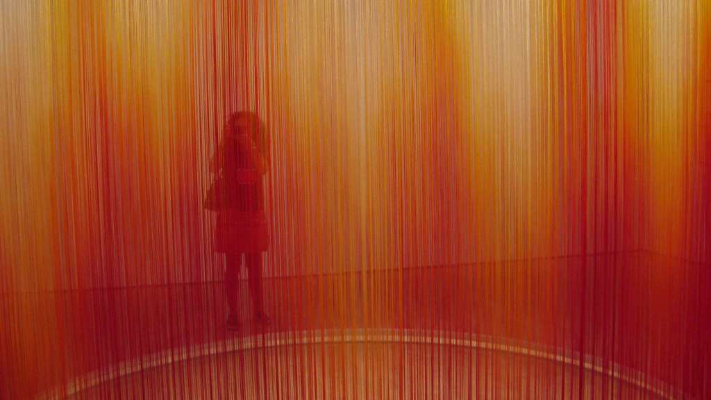 A person's silhouette is obscured by red, orange, and yellow strings stretched taut from the ceiling to the floor.