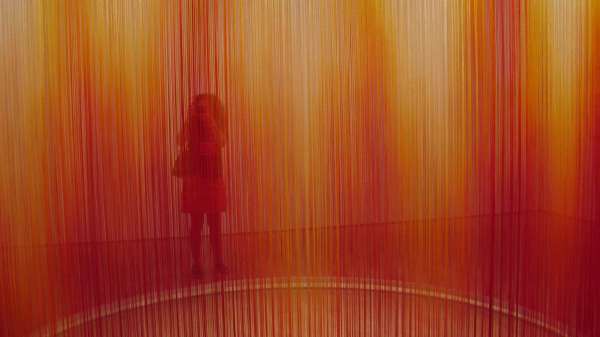 A person's silhouette is obscured by red, orange, and yellow strings stretched taut from the ceiling to the floor. 