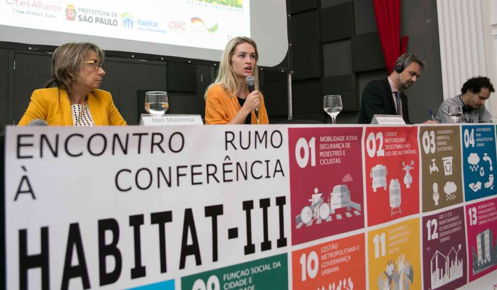 Speakers sit on stage for a panel at a table behind a colorful sign that reads: Encontro rumo à conferência Habitat III.
