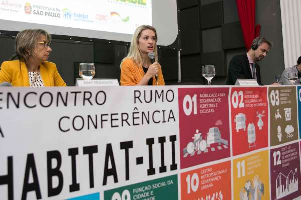 Speakers sit on stage for a panel at a table behind a colorful sign that reads: Encontro rumo à conferência Habitat III. 