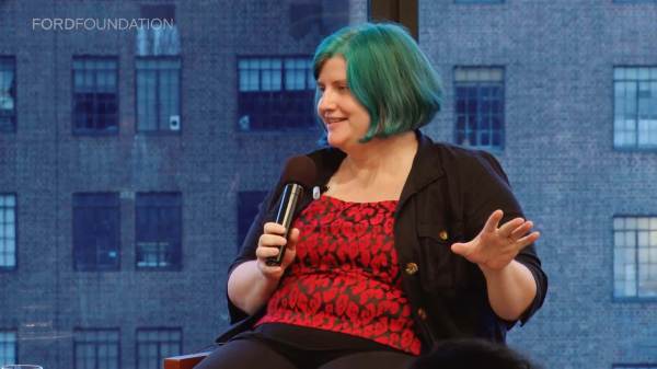 Cathy O'Neil sits on stage holding a mic. Her hair is in a bright teal bob. She wears a red and black patterned shirt with a black over shirt.