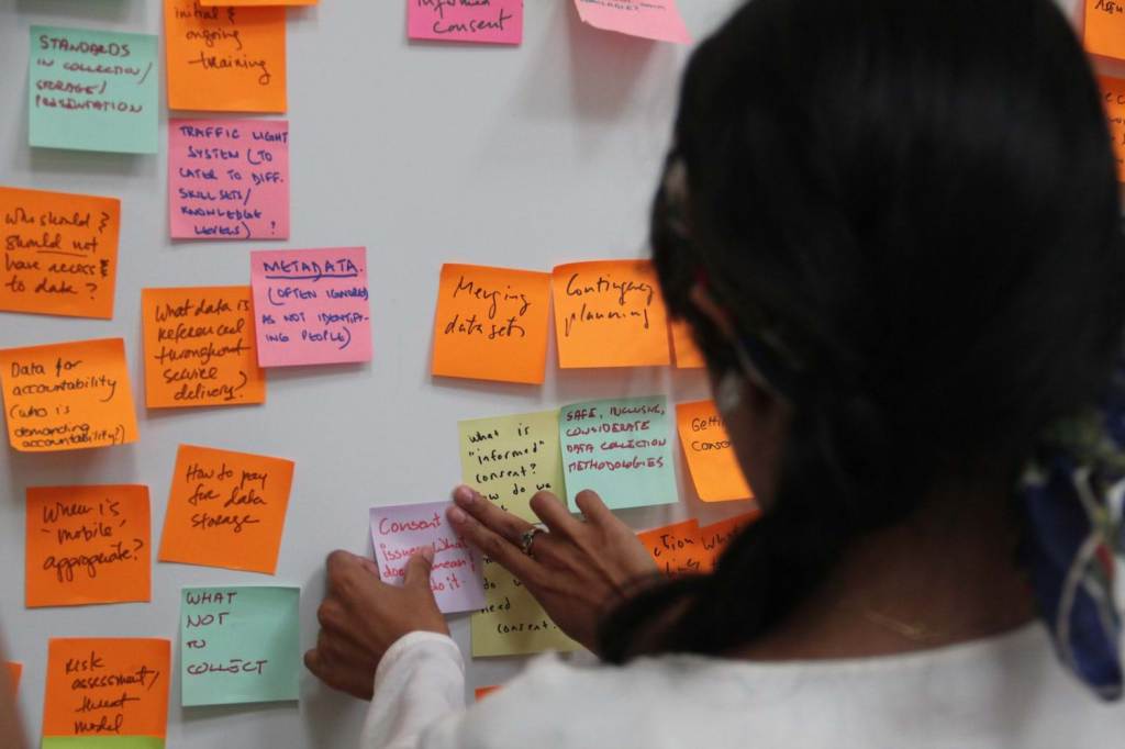 A person with dark hair stands in front of a wall covered in handwritten sticky notes about data collection and storage.