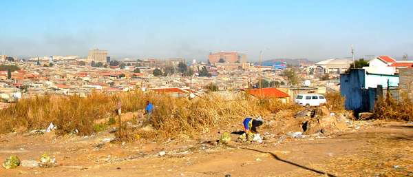 Black community members walk through the brush along the side of a dirt road. One stoops over a pile of refuse. Behind the long grass is a view of houses in Johannesburg.