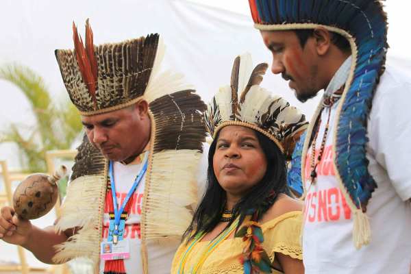 Sonia Bone Guajajara stands between two indigenous community members. They all wear indigenous headwear. Guadalajara's dark hair hangs below her shoulders and she wears a yellow shirt.