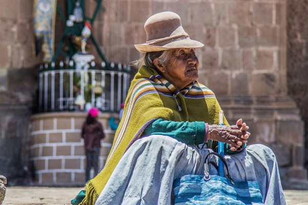A seated elderly woman, with a weathered face and hands, wears a light brown hat and a striped green and yellow poncho. A blue striped bag rests in her lap, and a blurred background shows stone architecture.