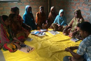 A group of seven women in colorful headscarves and traditional clothing are seated in a semicircle on a yellow tarp, facing a man. They are gathered in a room with exposed brick walls and papers scattered on the tarp.
