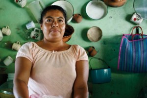 A medium shot of a woman with dark hair, wearing a light pink shirt, standing in a kitchen. The background shows a light green wall with various dishes and a blue striped bag hanging on it.