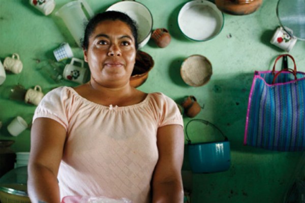 A medium shot of a woman with dark hair, wearing a light pink shirt, standing in a kitchen. The background shows a light green wall with various dishes and a blue striped bag hanging on it.