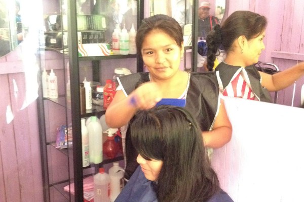 A smiling woman with dark hair, wearing a black smock, is styling the hair of a customer whose back is to the camera. In the background, another person with dark hair is visible in a mirror.