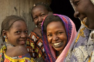A candid close-up of four women and children smiling, with one woman in the foreground looking at the camera. They wear colorful clothing and head coverings.