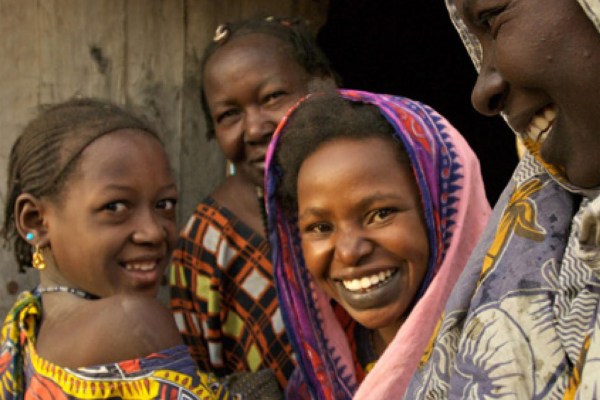 A candid close-up of four women and children smiling, with one woman in the foreground looking at the camera. They wear colorful clothing and head coverings.