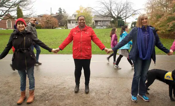 Three people in winter clothes hold hands and smile at the camera. Behind them, other people in winter clothes and sneakers walk across the street with a grassy field and suburban homes in the background.