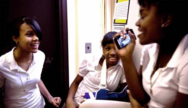 Three Black students with dark hair and bangs talk and laugh together. They are wearing white polo shirts and earrings. One has their arm in a cast with a blue and white sling. 