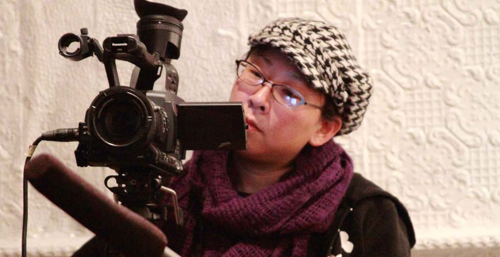 An Asian filmmaker wearing a houndstooth hat, glasses, and maroon scarf works behind a camera.