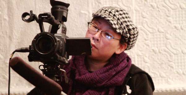 An Asian filmmaker wearing a houndstooth hat, glasses, and maroon scarf works behind a camera. 