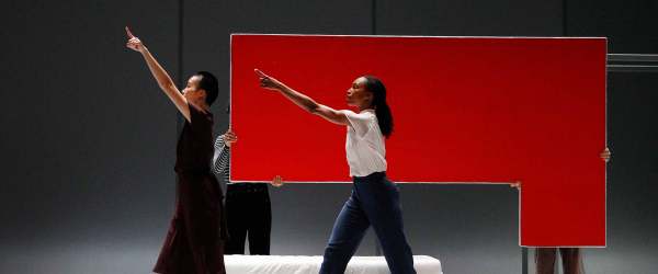 Two dancers dance face stage right, each with one arm stretched forward. Two people hold a large red shape behind the dancers.