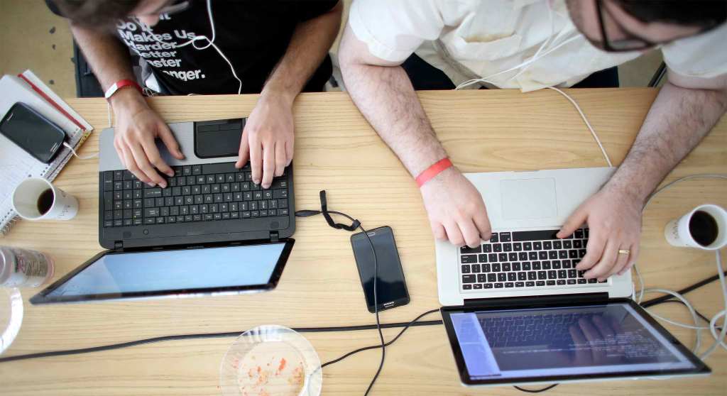 An overhead shot of a medium-skinned and light-skinned person typing on laptops set up on a shared wooden desk. Smartphones, cups of coffee, and a notebook rest on the desk.