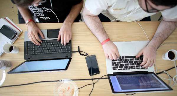 An overhead shot of a medium-skinned and light-skinned person typing on laptops set up on a shared wooden desk. Smartphones, cups of coffee, and a notebook rest on the desk.