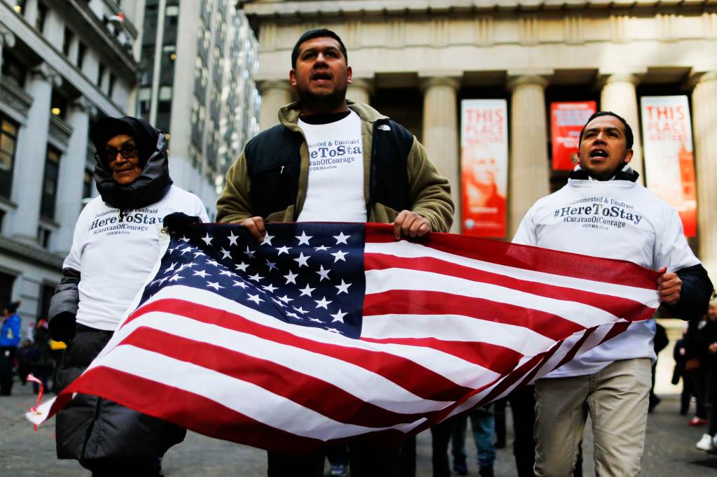 Three medium-dark skinned people march protest outside a buiilding while holding a US flag in front of them. They all wear white t-shirts that read "Undocumented & Unafraid, #HereToStay, #CaravanOfCourage. @DRMAction".