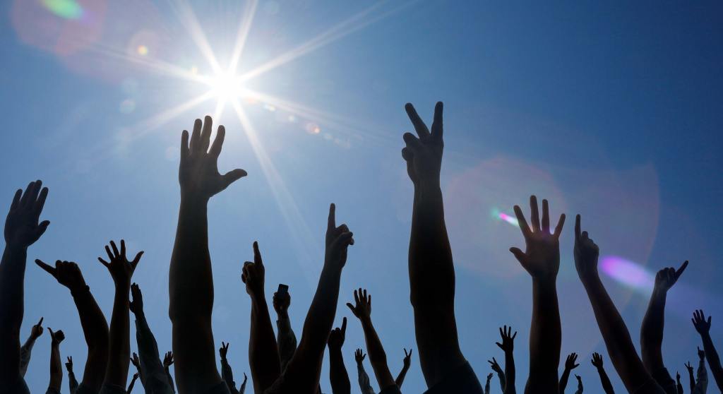 Many hands are raised to the sky, backlit by the sun to show only their silhouettes.