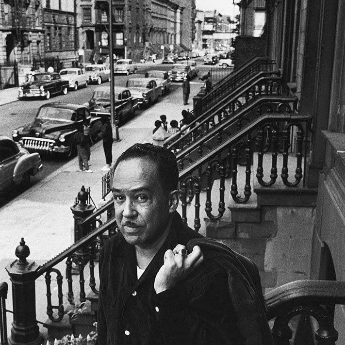 Black and white photo of Langston Hughes posing on the steps in front of his house in Harlem. He looks at the camera with a stoic expression, holding a jacket slung over his left shoulder. Brownstone stoops and the street are in the background.