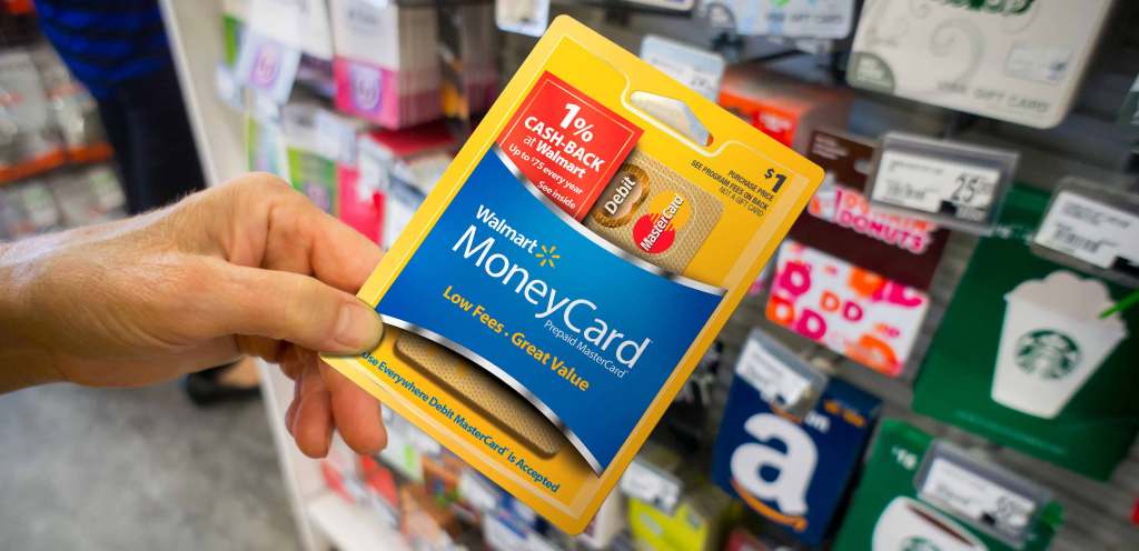 Close up of a person with a medium skin tone holding a pre-paid MoneyCard in front of a wall of gift cards and pre-paid cards.