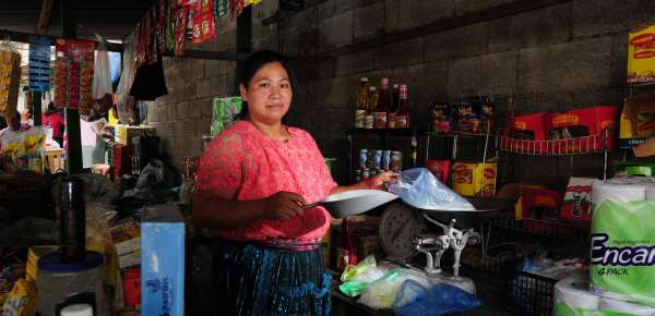 A Guatemalan person with medium skin tone stands in a market, prepared to scoop something into a bag on a scale. They wear a pink embroidered top with embroidered flowers and sheer short sleeves with a dark blue braided skirt and their black hair tied up. They are surrounded by colorful cans and boxes of store goods on shelves and hanging from the ceiling.
