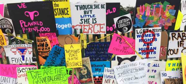 Colorful handmade signs from the Women's March hang from a fence. Some of the signs read: "Though she be little she is FIERCE", "A Woman's Place is in the Resistance", "LGBTQIA+ Equality", and "LGBT Muslim Women's Rights are Human Rights."