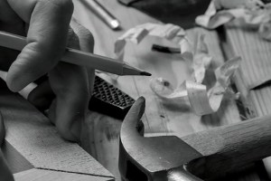 A black and white, close-up shot shows a hand holding a pencil over a piece of wood, with a hammer, wood shavings, and other tools scattered on a wooden surface in the background.