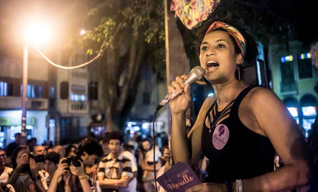 Marielle Franco speaks into a mic to a crowd outside at night. She wears a multicolored headband and black tank top with a large lavender sticker reading "Marielle:.