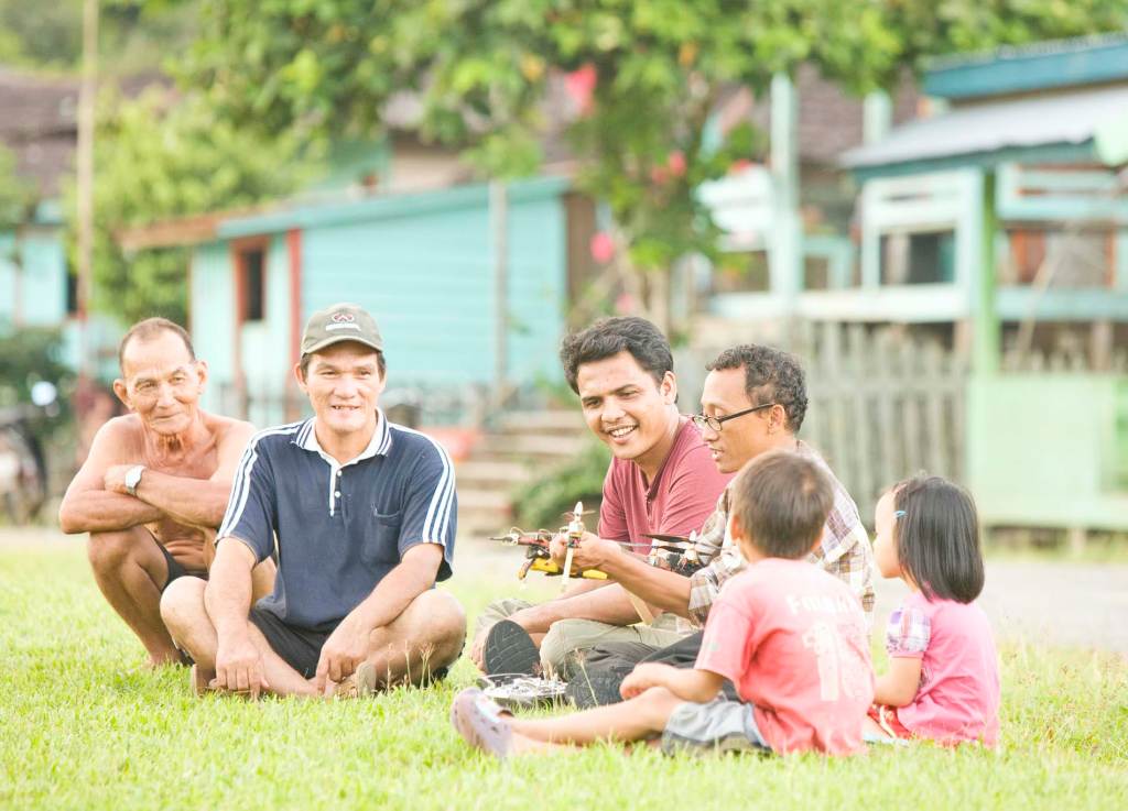 Six Dayak people of various ages, including two children, sit outside on the grass in Setulang. They smile, focused on a person in the middle holding a drone.