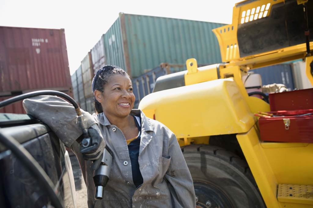 A female African American mechanic holds a drill, fixing machinery in industrial container yard.