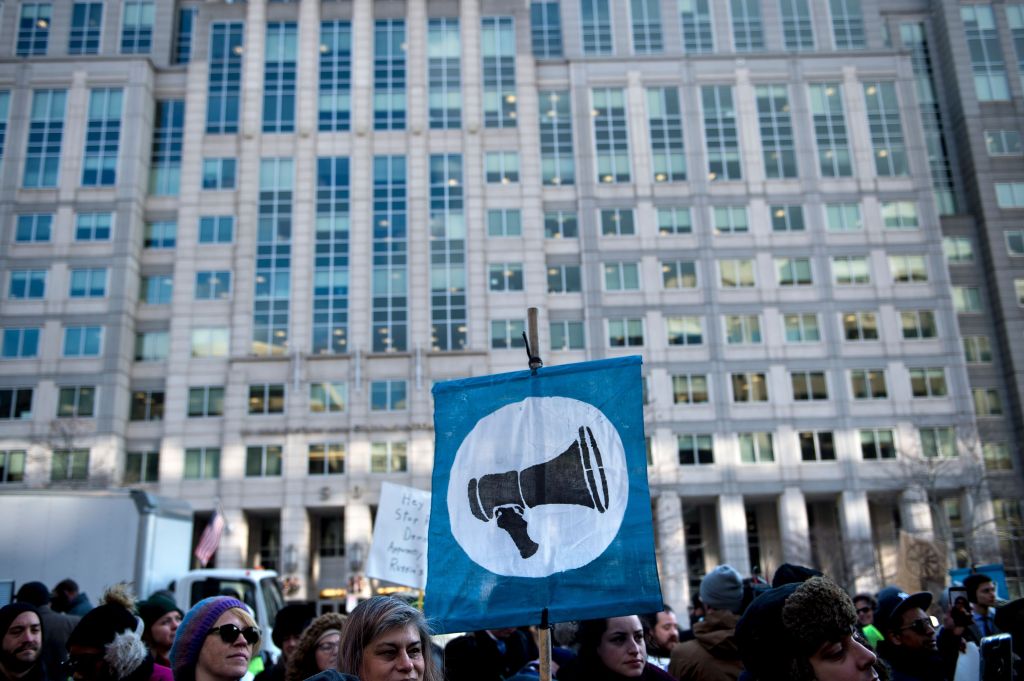 Protestors in front of building holding a blue banner with a bullhorn graphic on it.