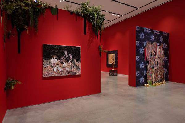 Interior view of the Ford Foundation Gallery featuring the "Radical Love" exhibition. A large, intricate tapestry made of colorful beads and sequins is displayed on a dark patterned wall, with vibrant red gallery walls and lush botanical installations visible in the background.