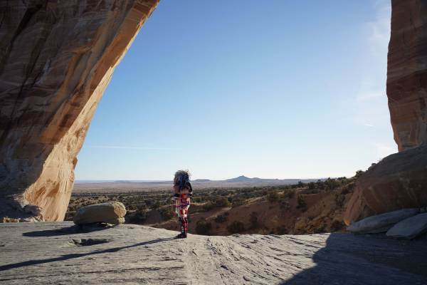 A photo of Hanska Luger's "The One Who Checks & The One Who Balances". A person wearing a colorful multi-patterned jumpsuit stands on a rock cliff looking out onto a desert with patches of green brush dotting the landscape.