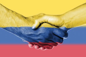 A close-up image of two hands shaking, painted with the colors of the Colombian flag: yellow on top, blue in the middle, and red at the bottom, symbolizing peace and unity.