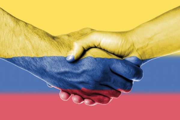 A close-up image of two hands shaking, painted with the colors of the Colombian flag: yellow on top, blue in the middle, and red at the bottom, symbolizing peace and unity.