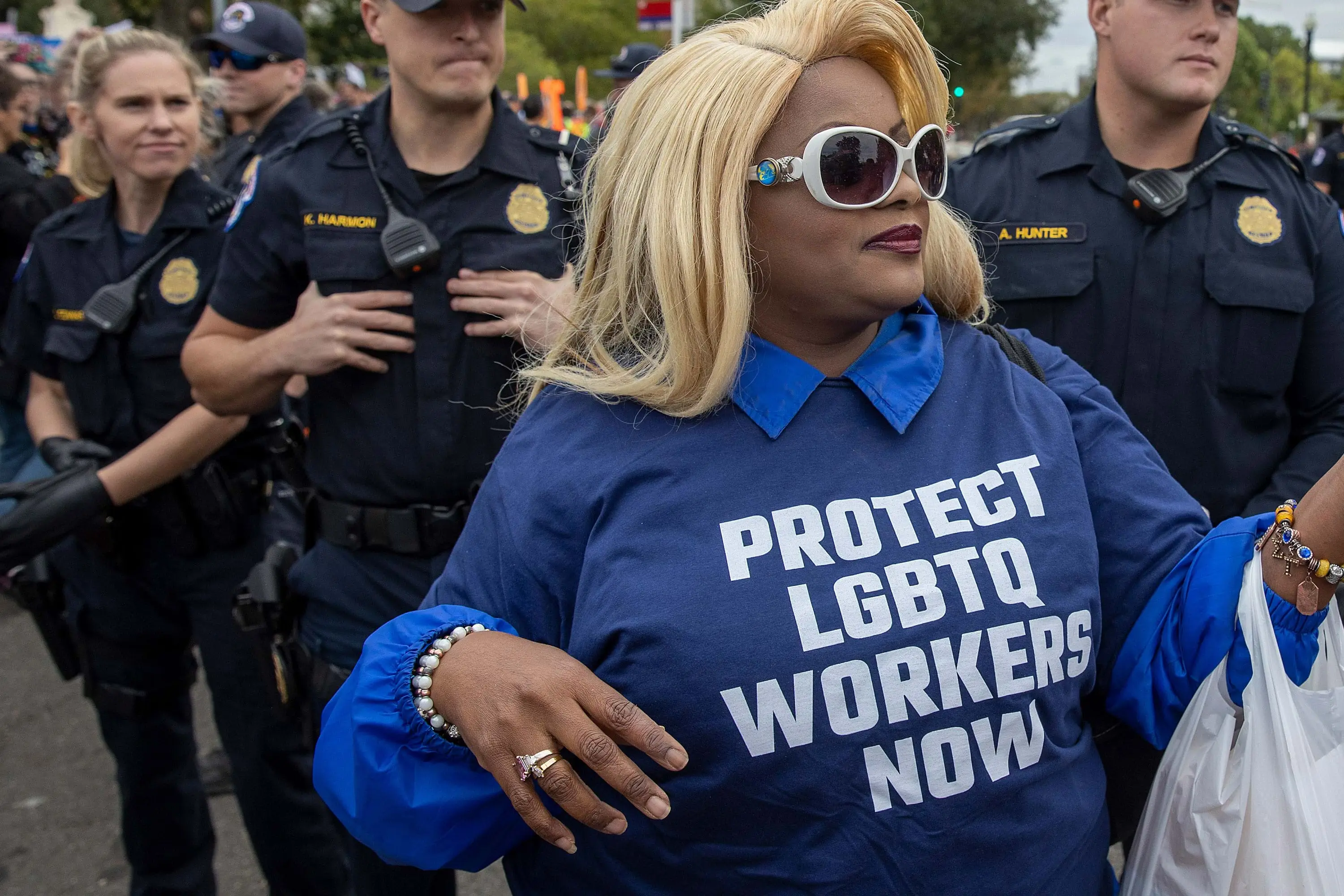 A Black person in sunglasses, long blonde hair, and a shirt that says "PROTECT LGBTQ WORKERS NOW" stands with their back to white police officers.