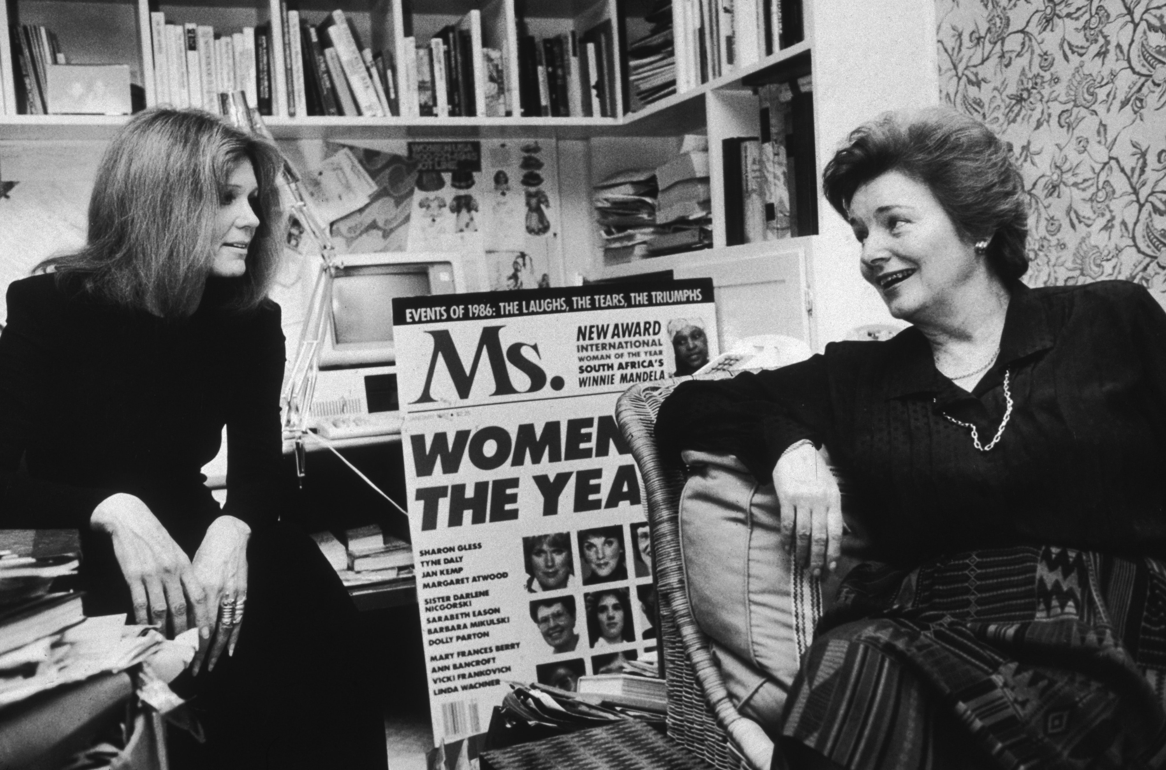 A black and white photo of Gloria Steinem sitting with another woman in an office space with a large display of a Ms. magazine cover of the Women of the Year issue.
