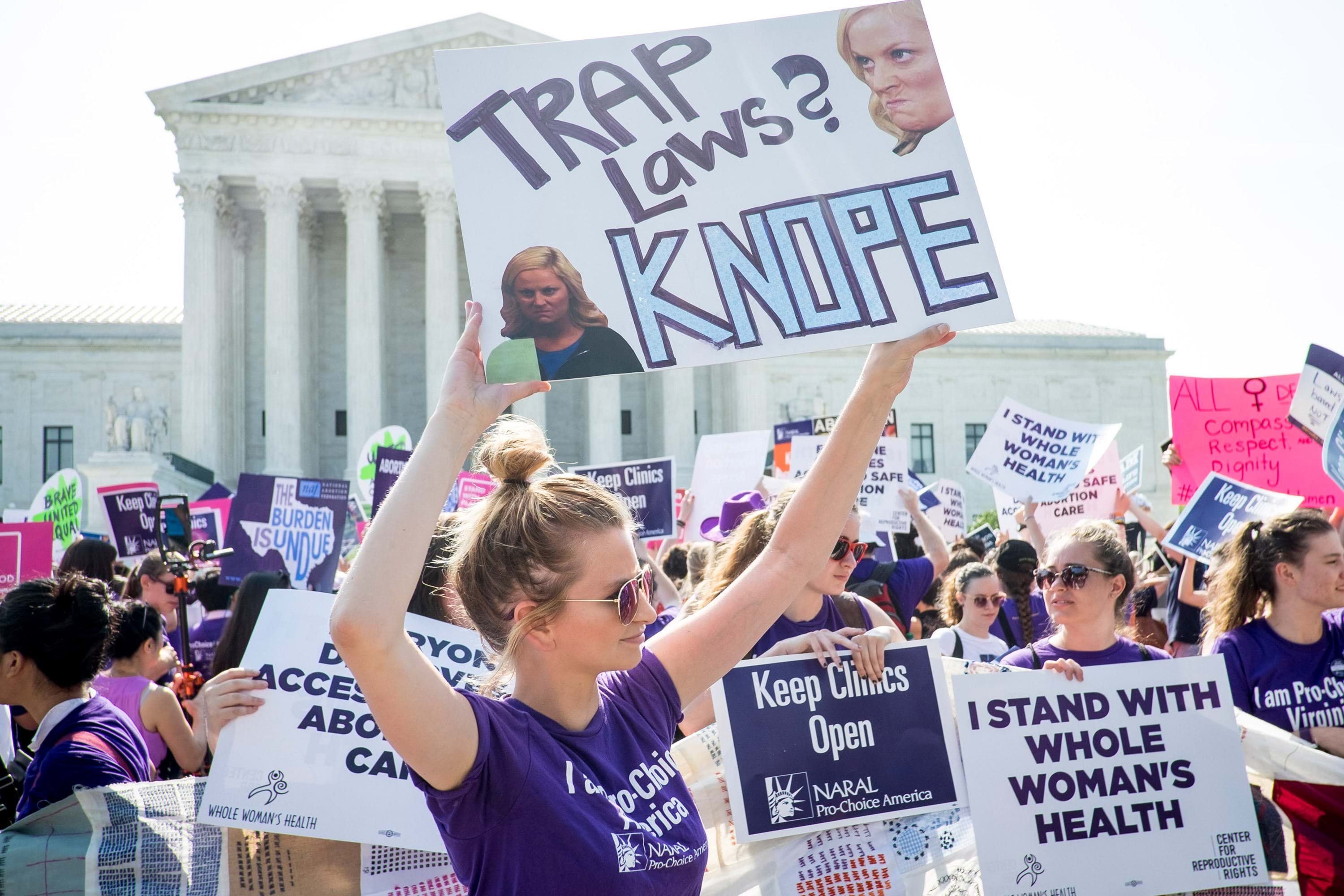 A white person in a purple t-shirt with their hair in a top knot  protests with a large crowd in from of the Supreme Court. They hold a sign above their heads that says "TRAP Laws? KNOPE" with photos of Amy Poehler as Leslie Knope. 