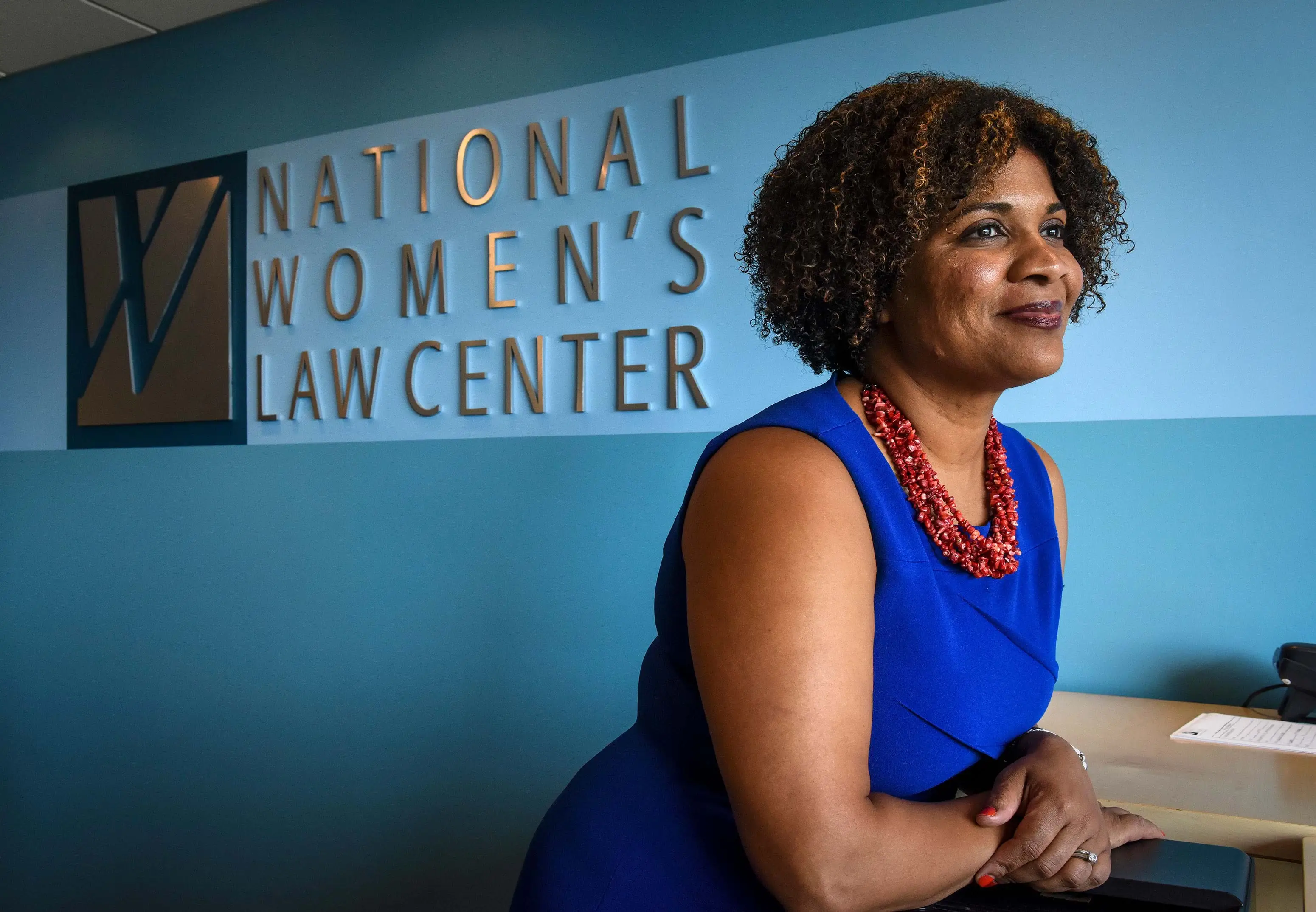 Fatima Goss Graves, a Black woman wearing a bright blue dress and coral necklace, stands in front of a large wall sign for the National Women's Law Center.