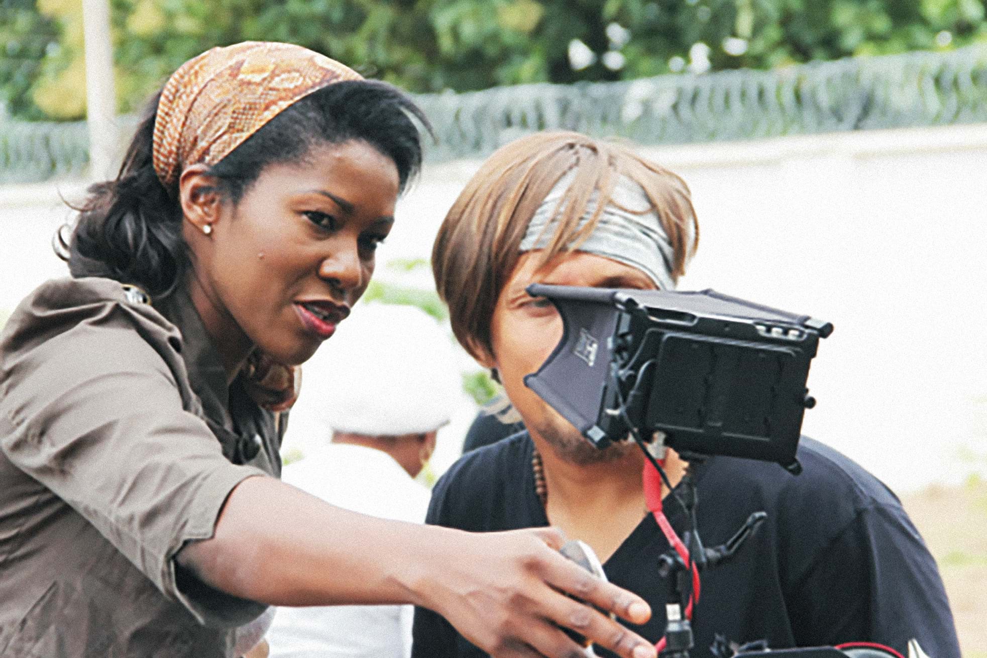 Stephanie Okereke, a Black woman wearing a brown bandana over her shoulder length hair, directs a cinematographer as they look through 