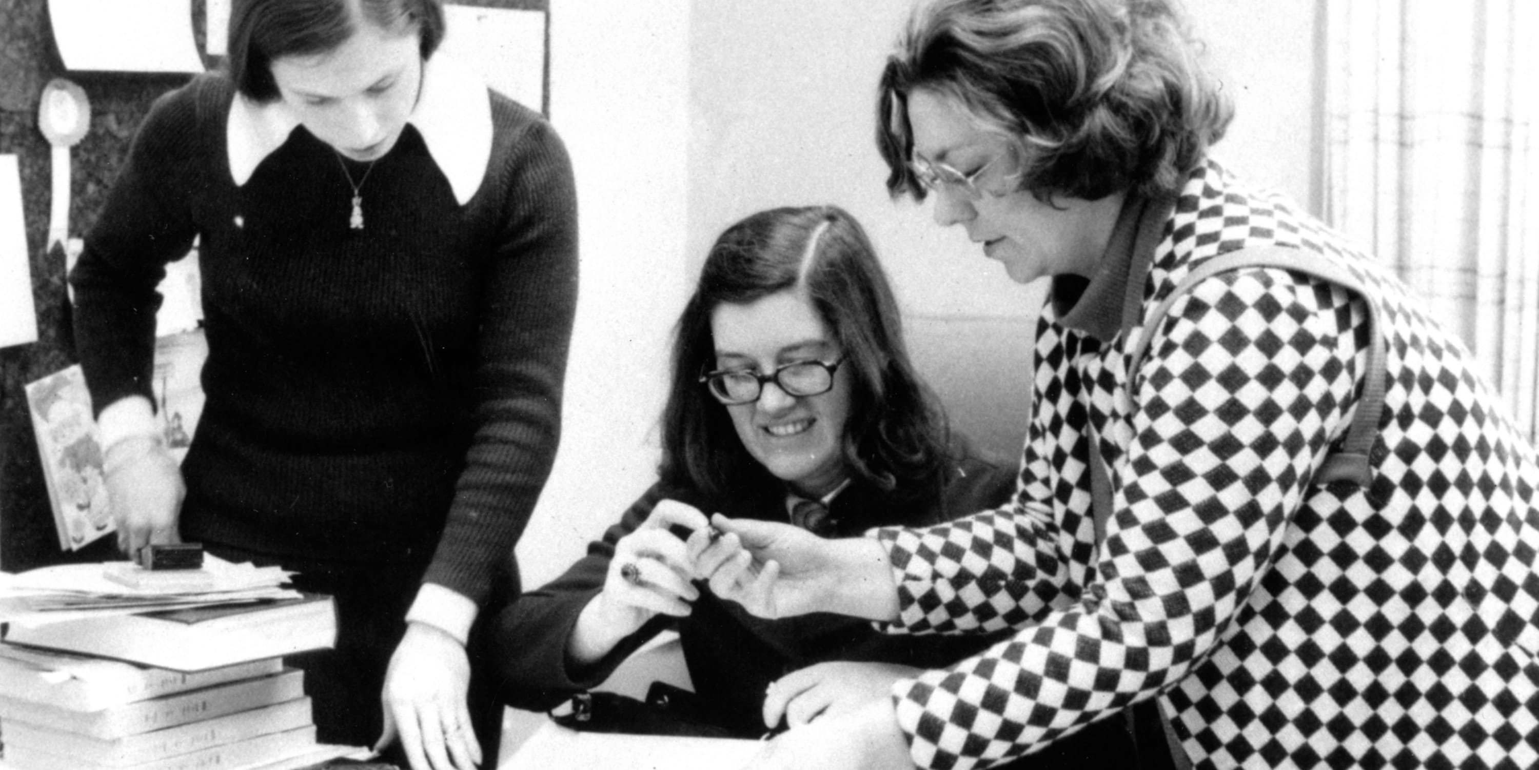 A black and white photo of three white women talking together at a desk next to a stack of books.