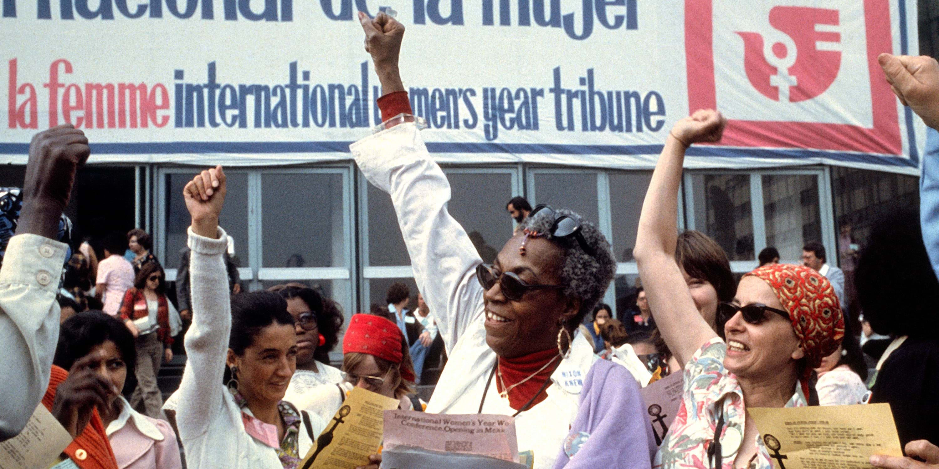 A photo of a crowd of people, including a Black woman in the foreground, with their fists raised in the air at a protest. 