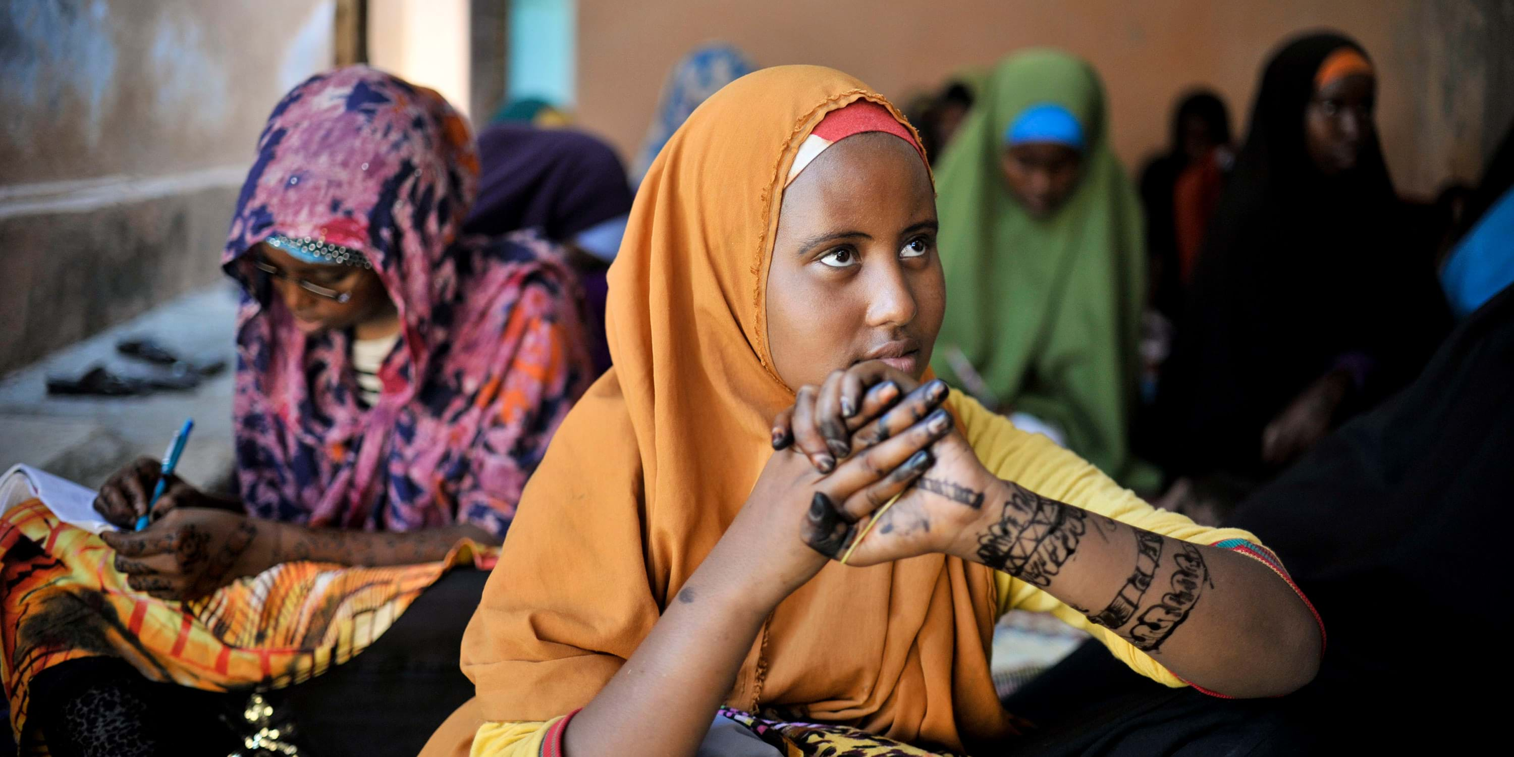 A group of women with different shades of brown skin wearing different colored hijabs focused on reading or writing on papers balanced on their laps. A woman with a bright orange hijab and black drawings on her arms looks up. 