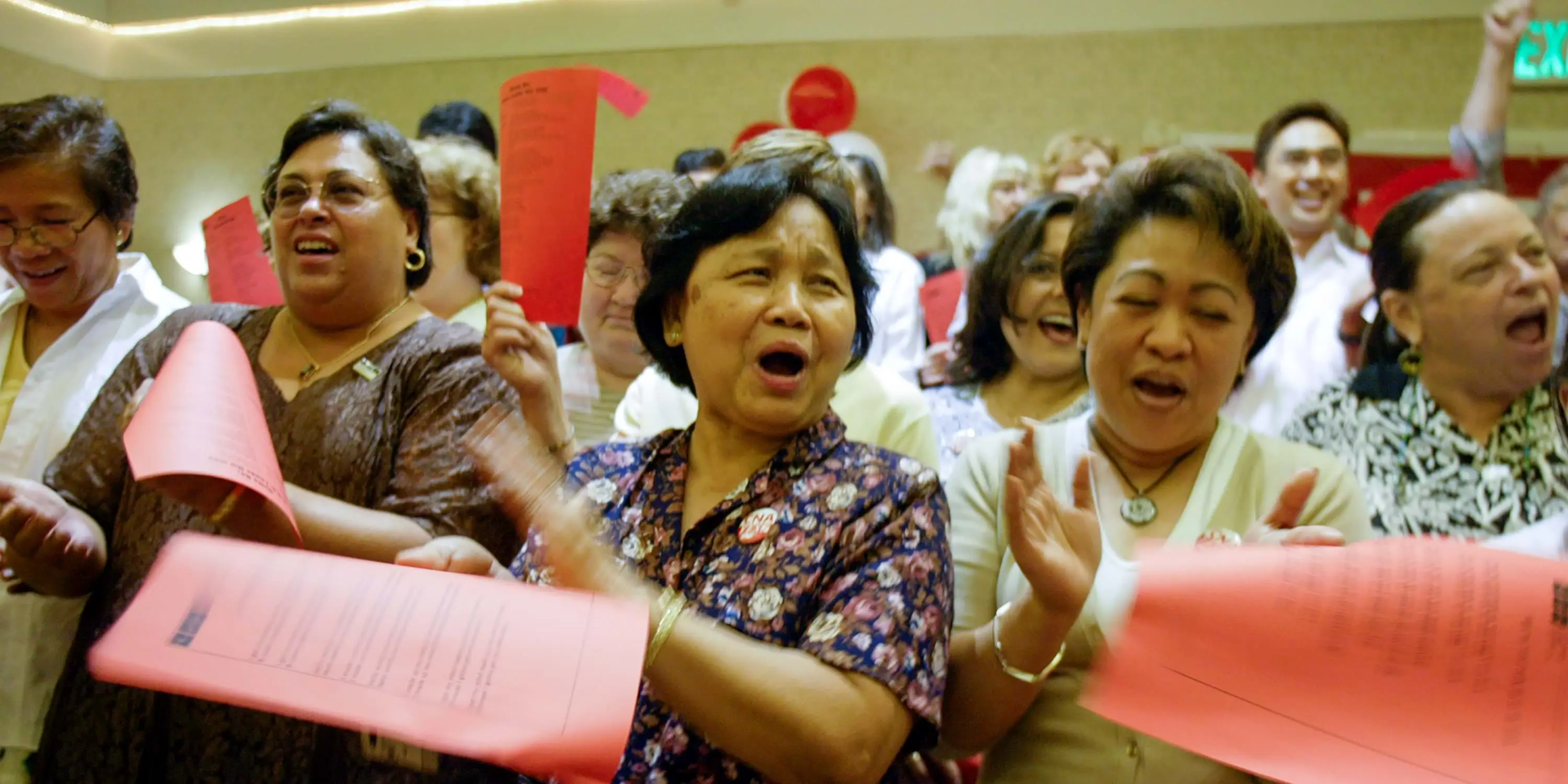 A row of Asian women carrying red pieces of paper cheer and applaud in a crowd.
